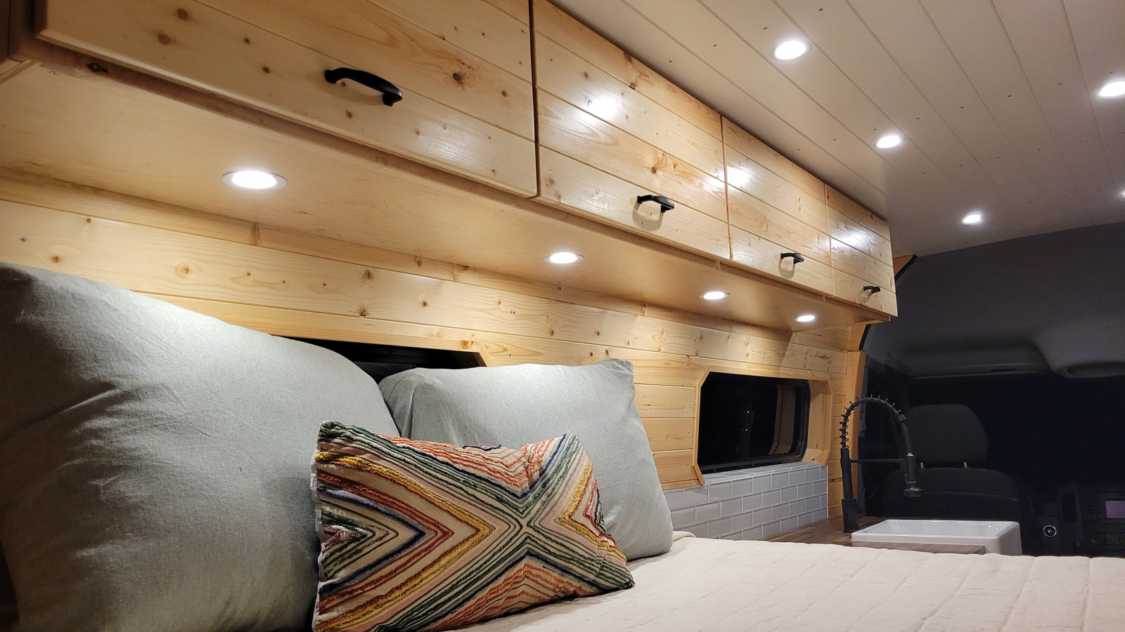 Inside a bright RV rental: wooden walls, overhead cabinets, a cozy bed, and a compact sink. Ideal mobile living.