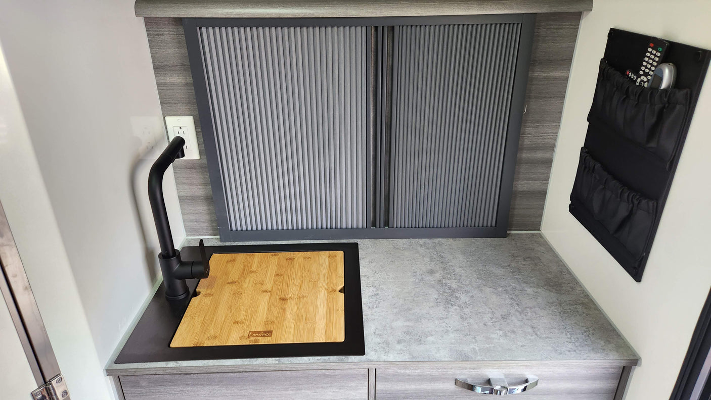 Compact RV rental kitchen counter with sleek black faucet, built-in sink/cutting board, and tambour storage unit.