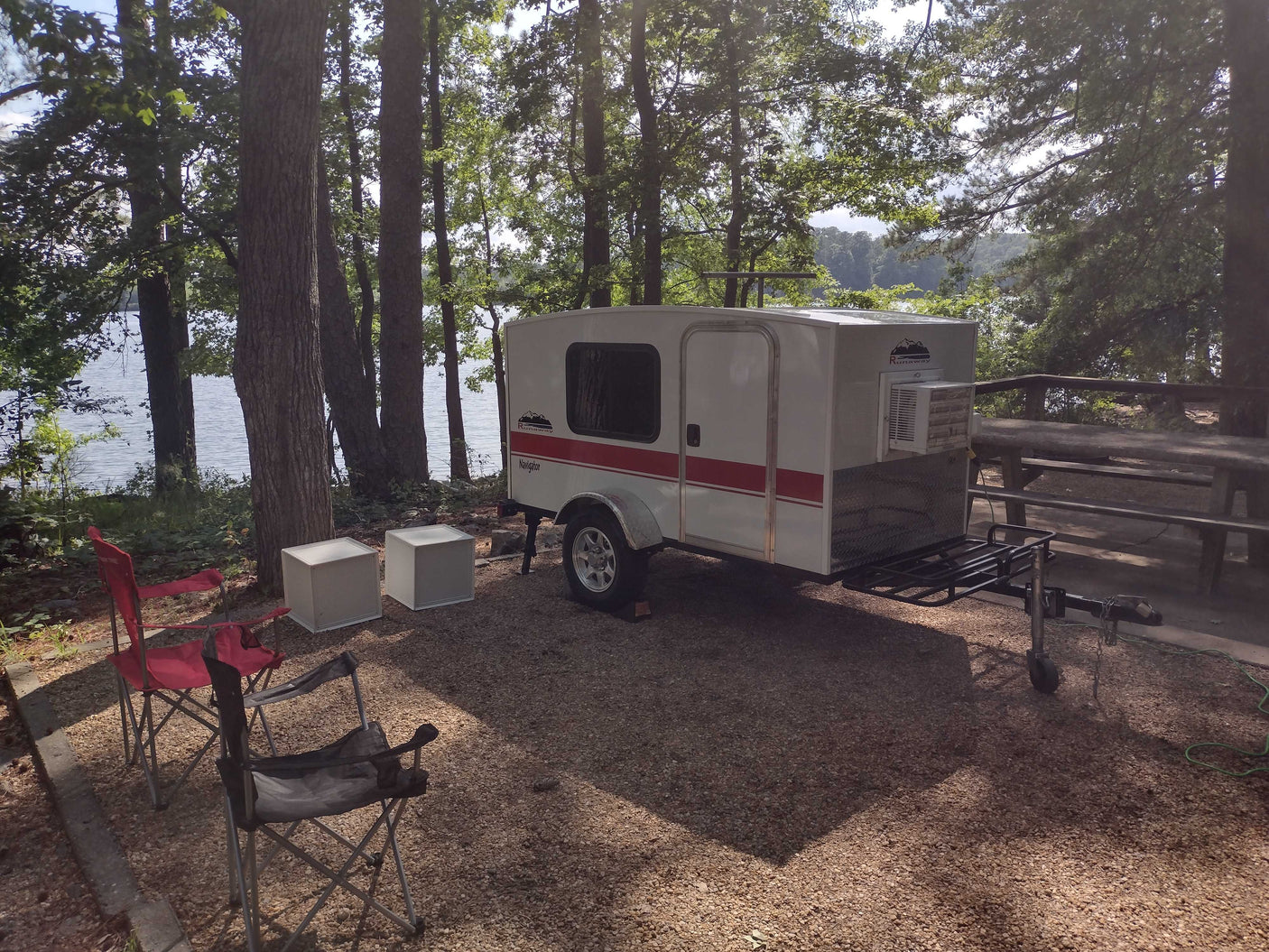 Compact teardrop RV rental at a scenic lakeside campsite under tall trees, ideal for outdoor adventures.