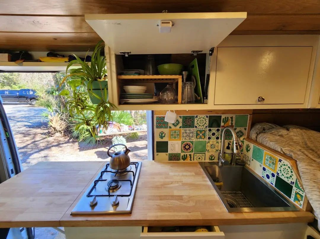 RV rental interior: a compact kitchen with wood counters, a stove, sink, and vibrant tiled backsplash. Ready for travel adventures.