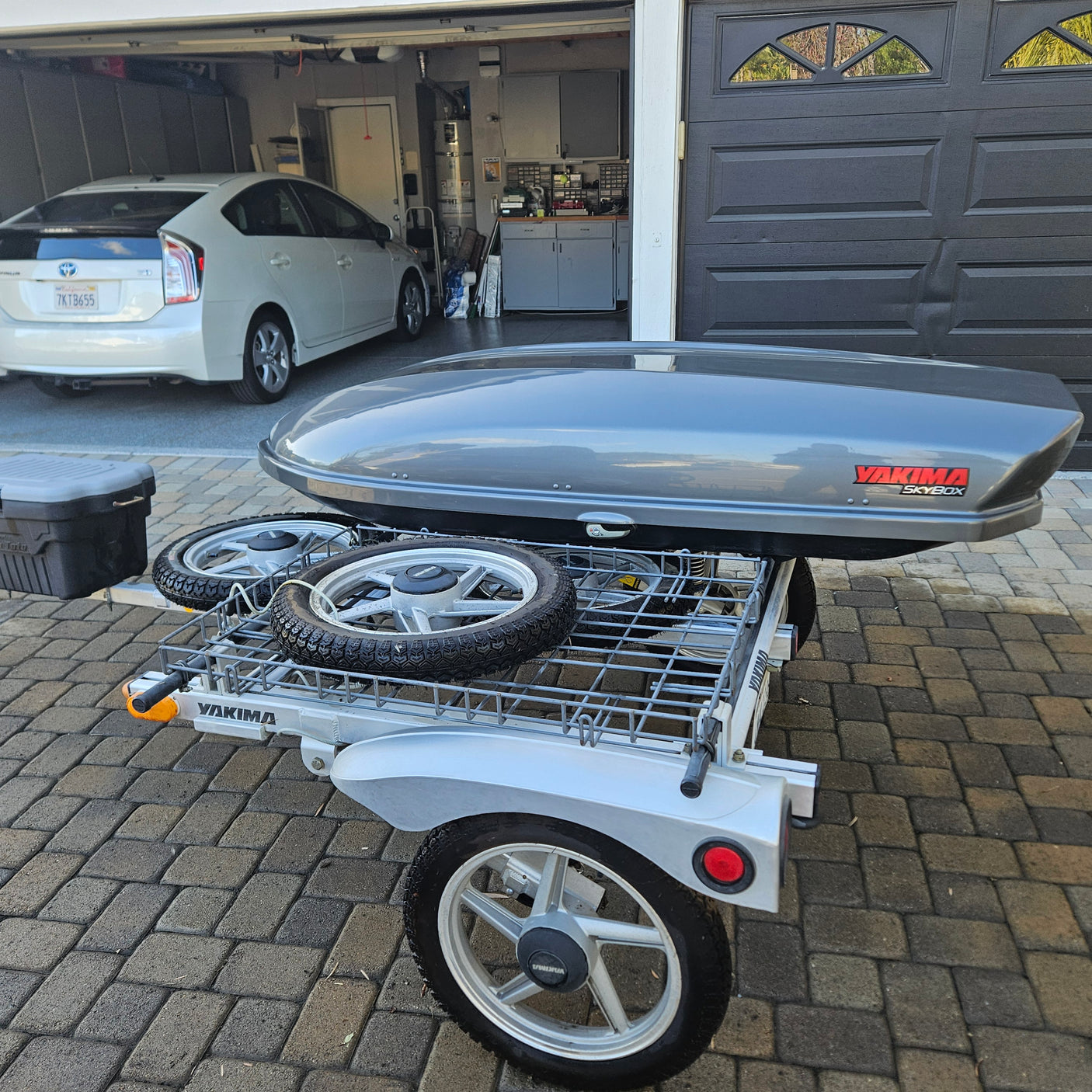 Grey Yakima utility trailer with SkyBox cargo box & spare tires, parked on pavers. Ideal for RV rental trips/gear hauling.
