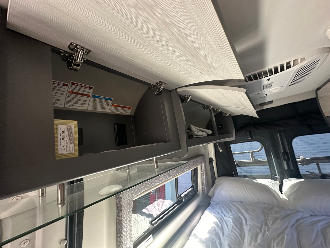 Open overhead storage cabinets in an RV rental, showing a cribbage set. A ceiling AC unit and bed are also visible.