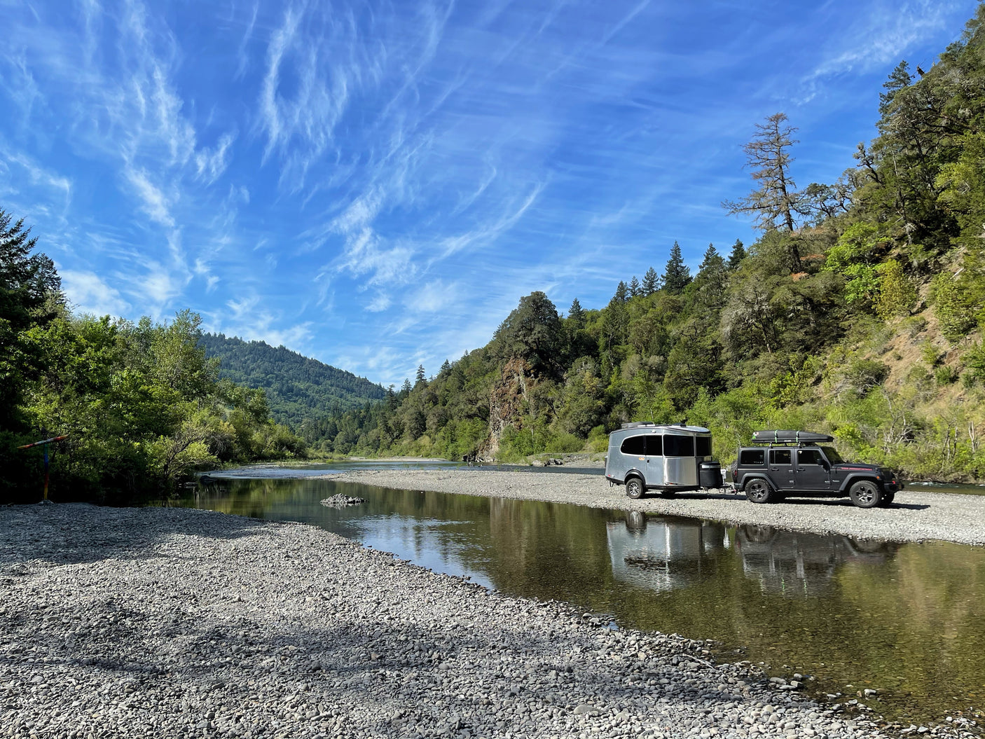RV rental: Grey trailer & black Jeep parked on a rocky riverbank, calm river reflecting green mountains under blue sky.