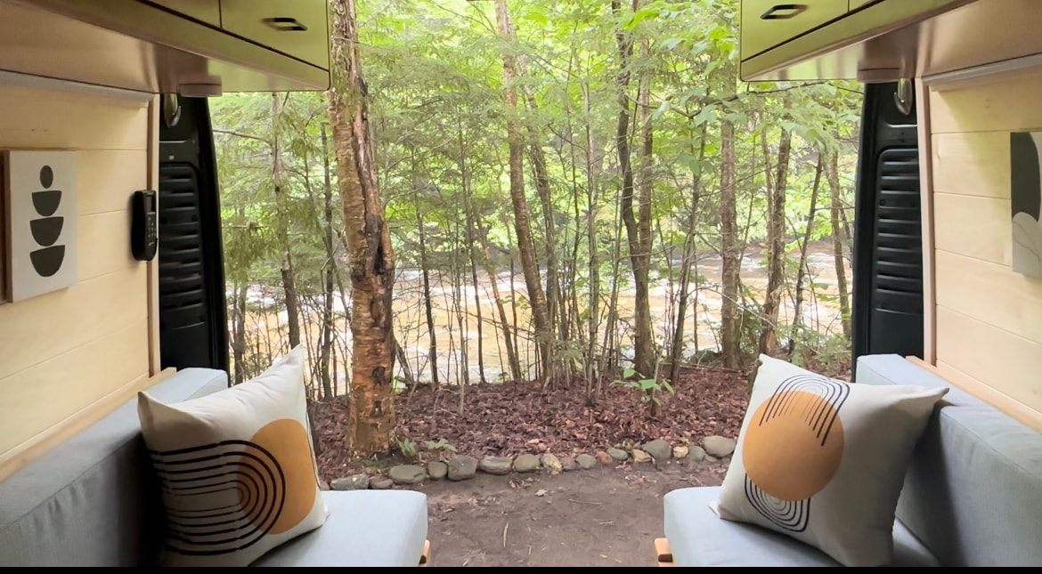 Cozy RV rental interior with open back doors, offering a serene view of a forest and flowing river. Perfect nature escape.