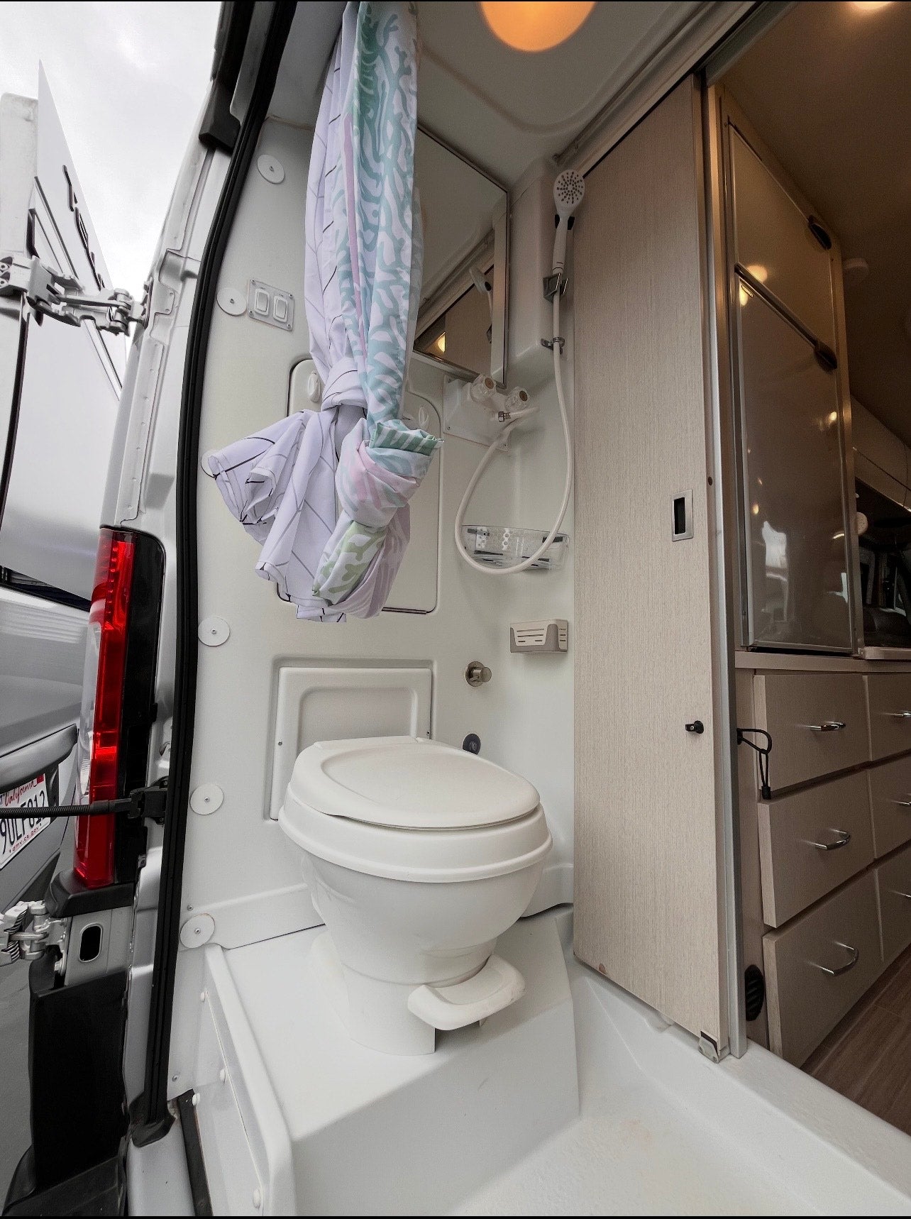 This compact rv rental bathroom has a white toilet, shower, mirror, and a tied-back curtain. Light cabinetry is on the right.