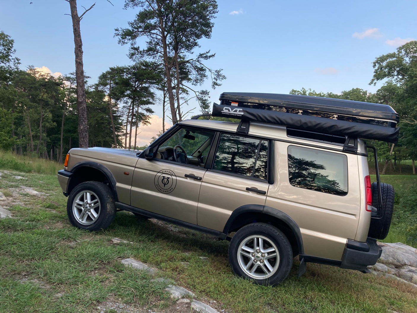 A tan Land Rover Discovery 2, equipped with a rooftop tent & awning, parked on a rocky trail. Ideal for adventure travel or an off-road RV rental.