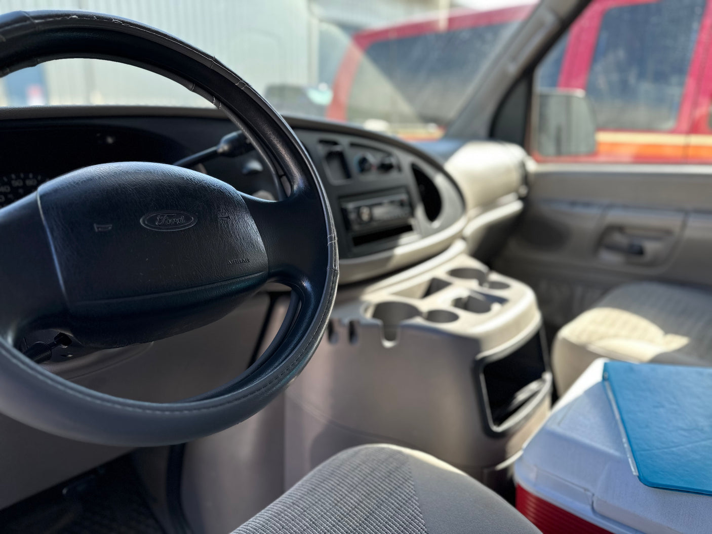 Driver's view inside a gray Ford van RV rental, showing the steering wheel, dashboard, and a red cooler.