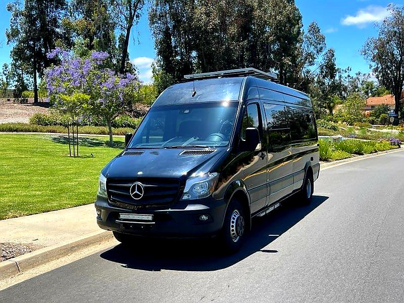 Black Mercedes Sprinter van, likely an RV rental, parked on a sunny street next to green grass and trees.