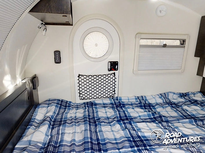 RV rental interior features a cozy bed with blue plaid comforter, round door, window, and wall-mounted controls.