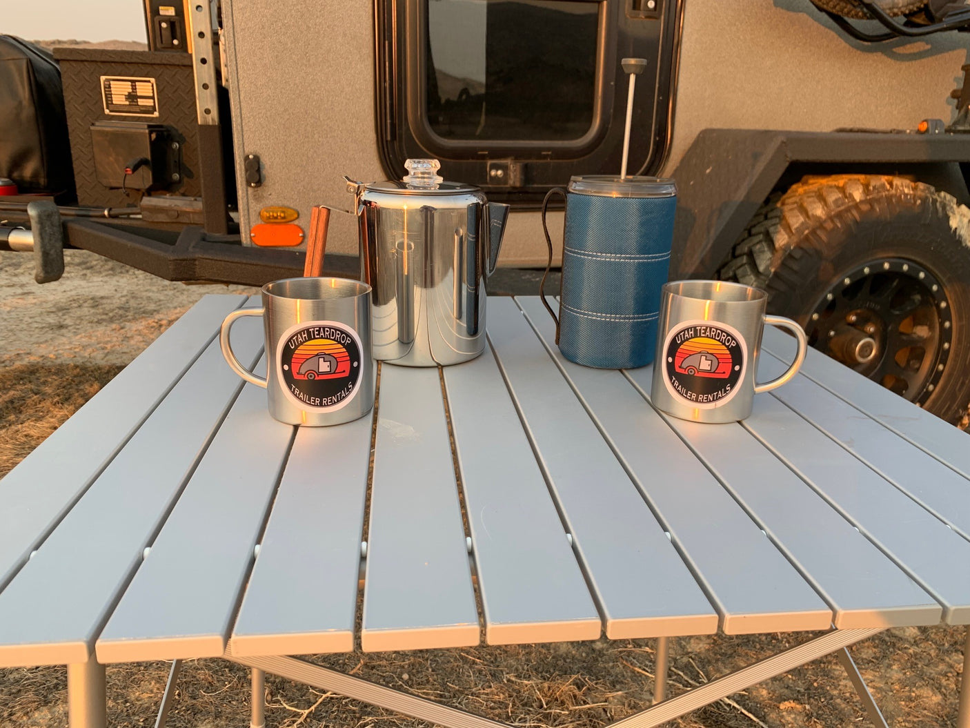 Morning coffee by your Utah Teardrop RV rental! Two custom mugs, percolator, & French press on a camp table.