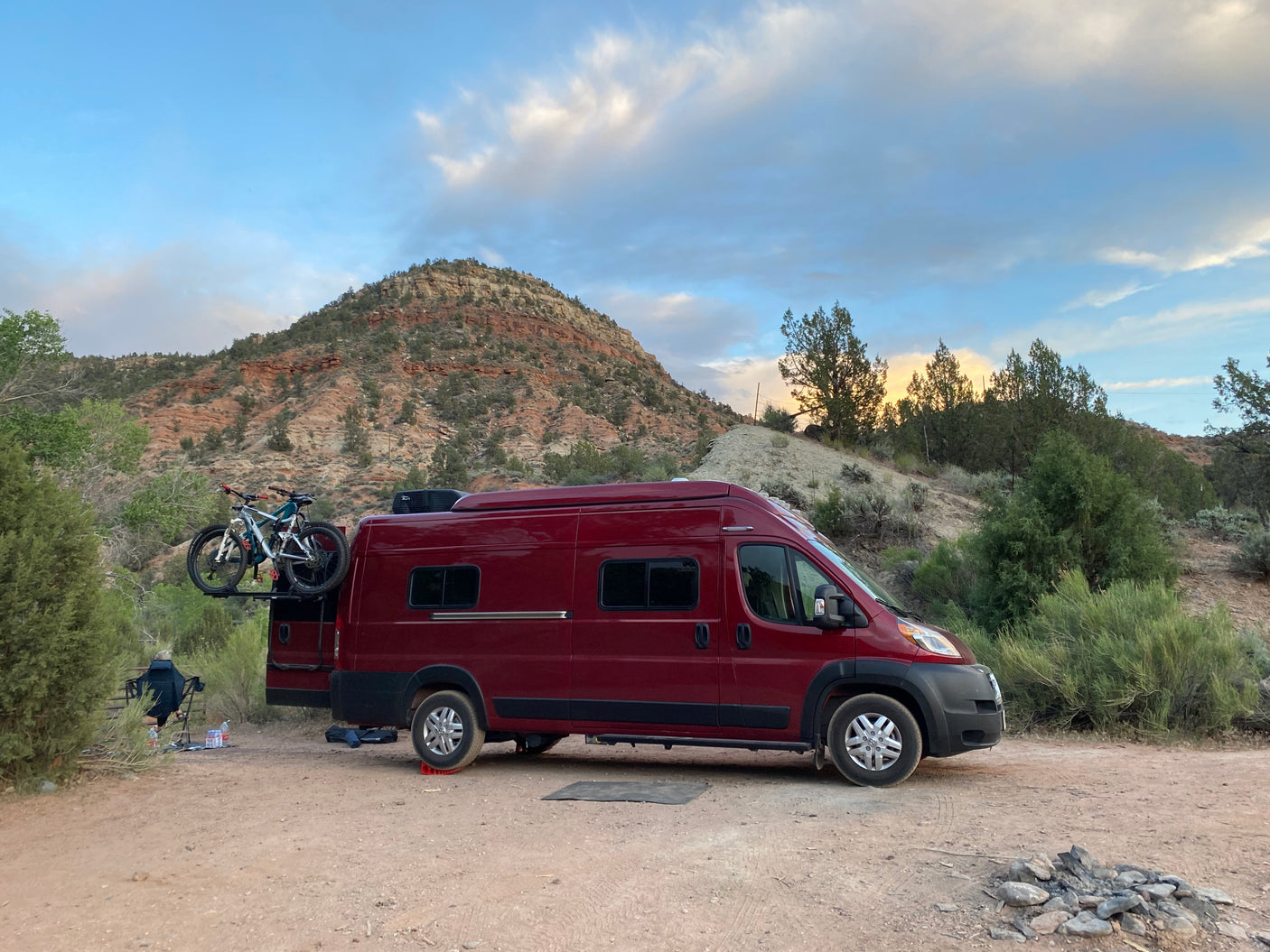 Scenic RV rental setup: red camper van with bikes, leveled for desert camping. Rocky mountains & sunset sky.