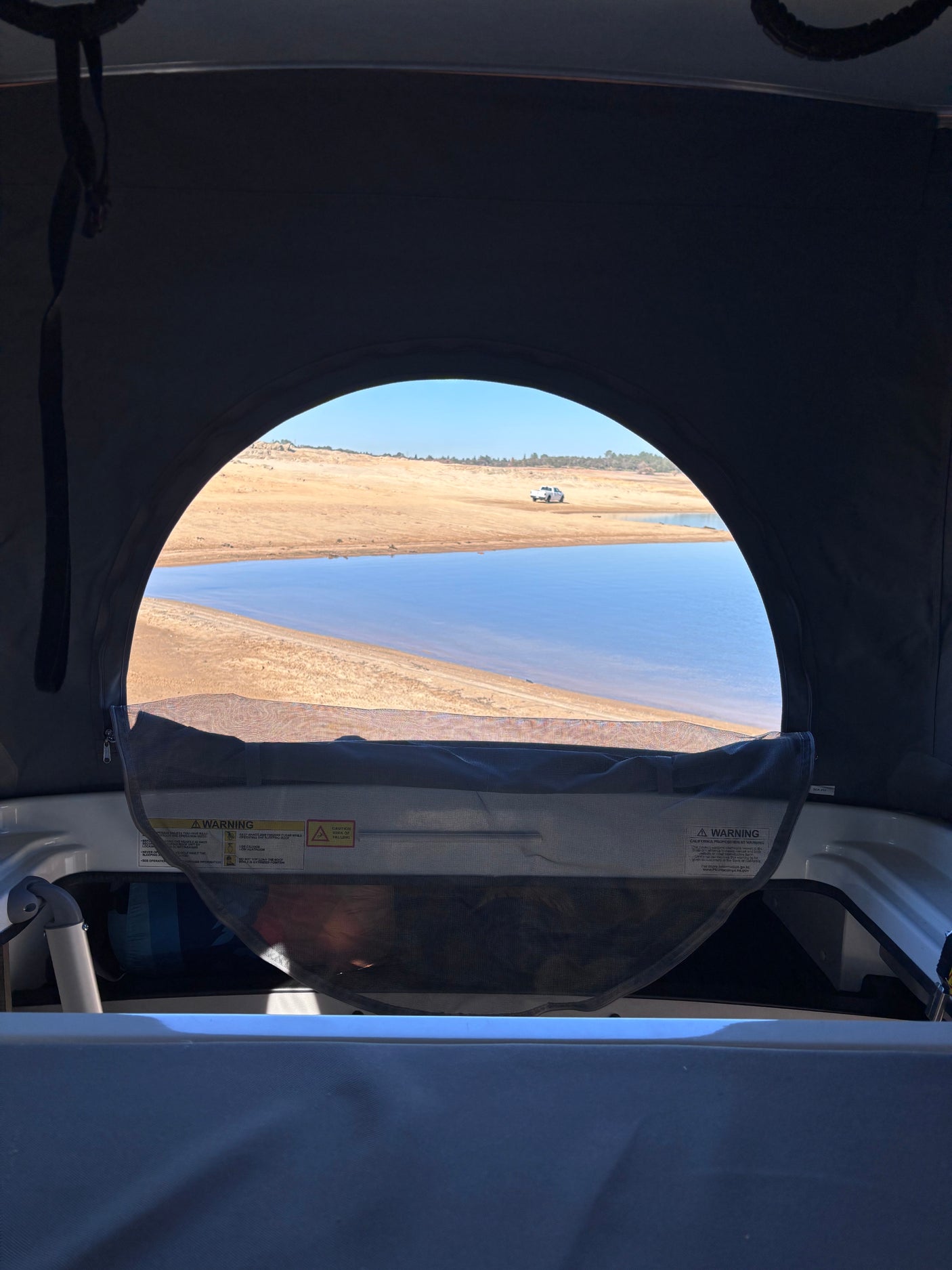 Inside an RV rental pop-top camper, a serene lake and sandy shore with a distant truck are framed by the window.