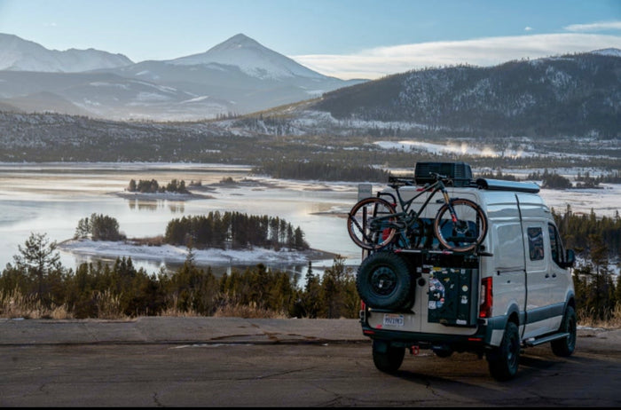 An RV rental camper van, equipped with bikes, overlooks a picturesque frozen lake and majestic snow-capped mountains.