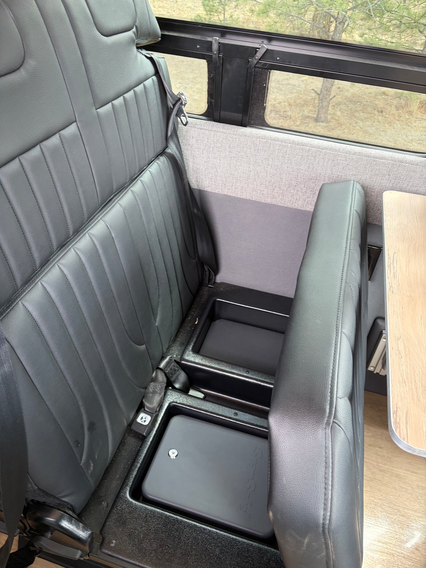 This RV rental interior features black passenger seats with seatbelts, under-seat storage bins, and a window with a nature view.