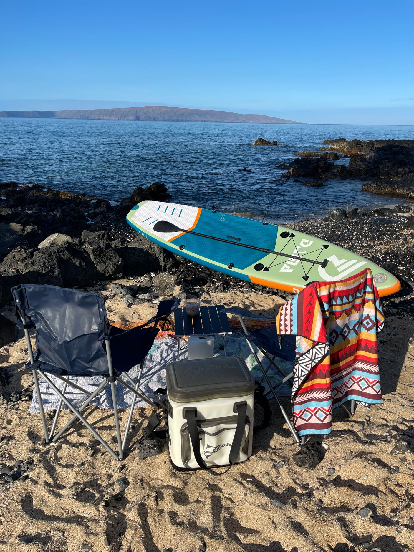 A tranquil beach scene: paddleboard, chairs, and cooler set up on sand and rocks by the ocean. An ideal RV rental getaway spot.
