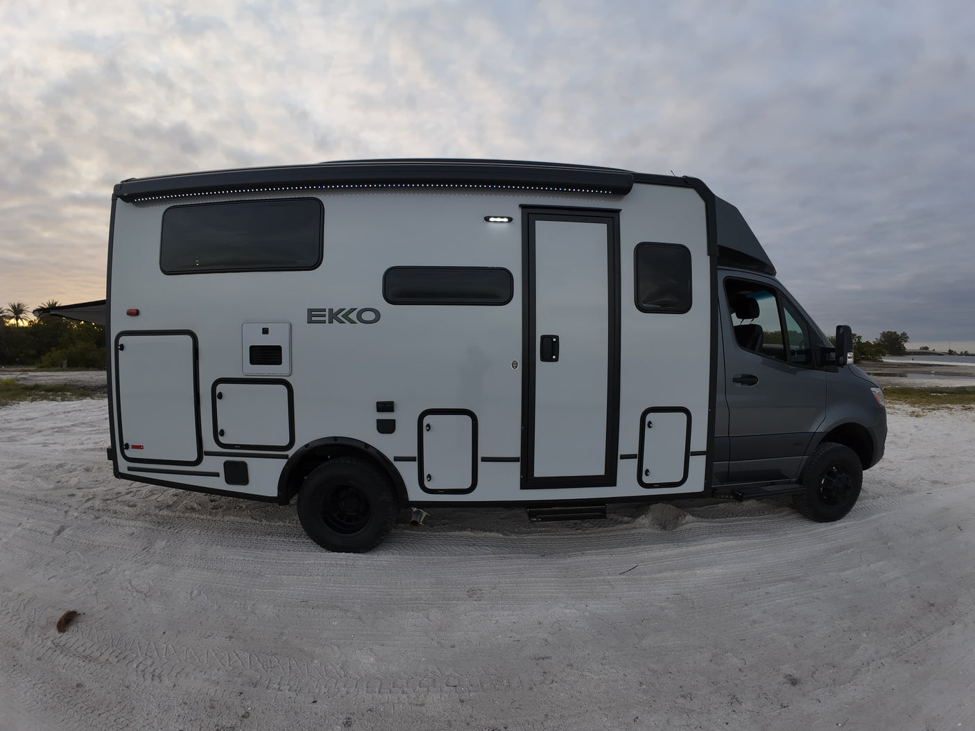Modern Winnebago EKKO RV rental parked on sandy ground by water under a cloudy sky, ideal for outdoor adventures.