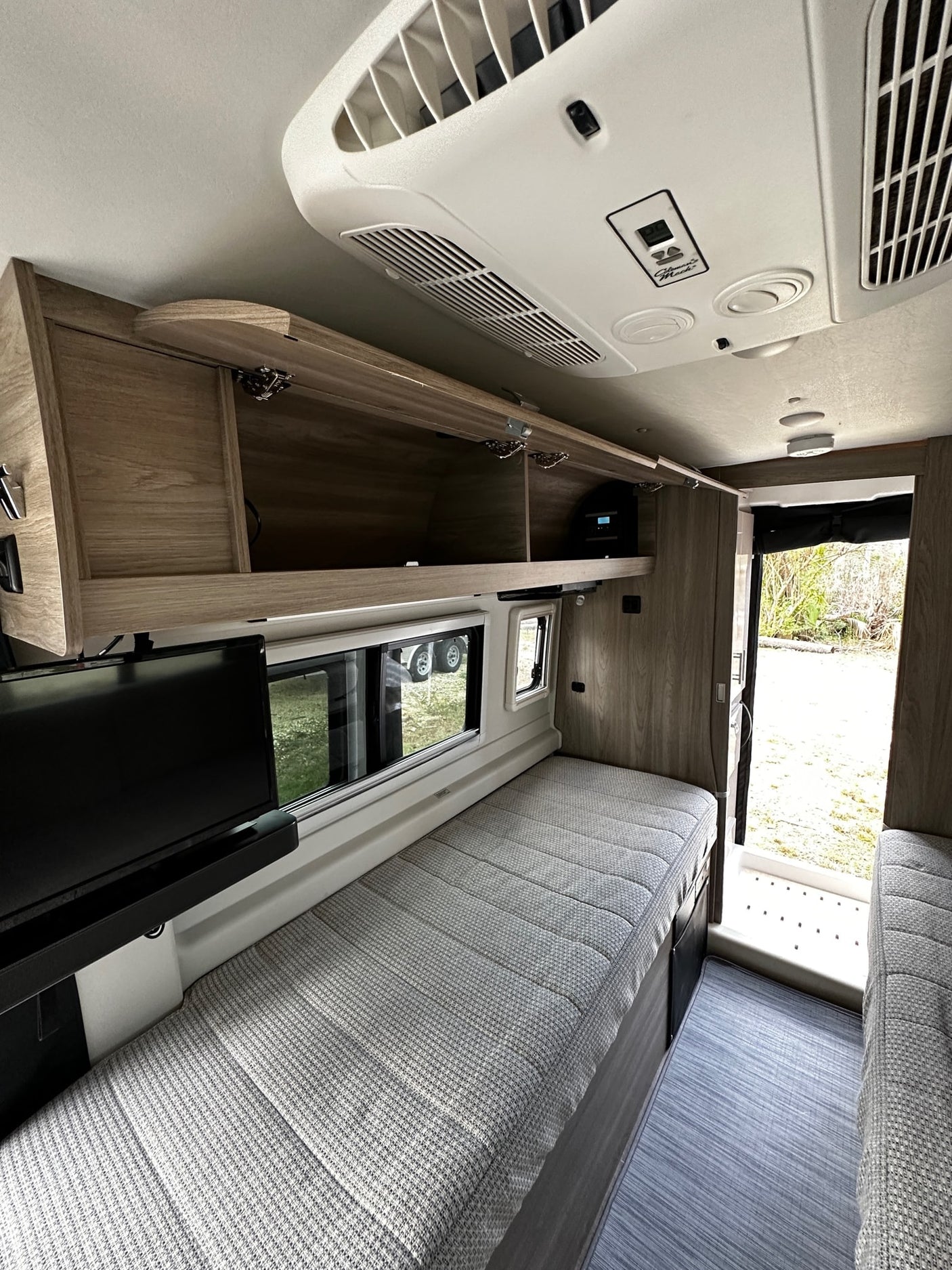 RV rental interior: Overhead AC unit, light wood cabinets, a long bed/seating area, and side windows providing natural light.