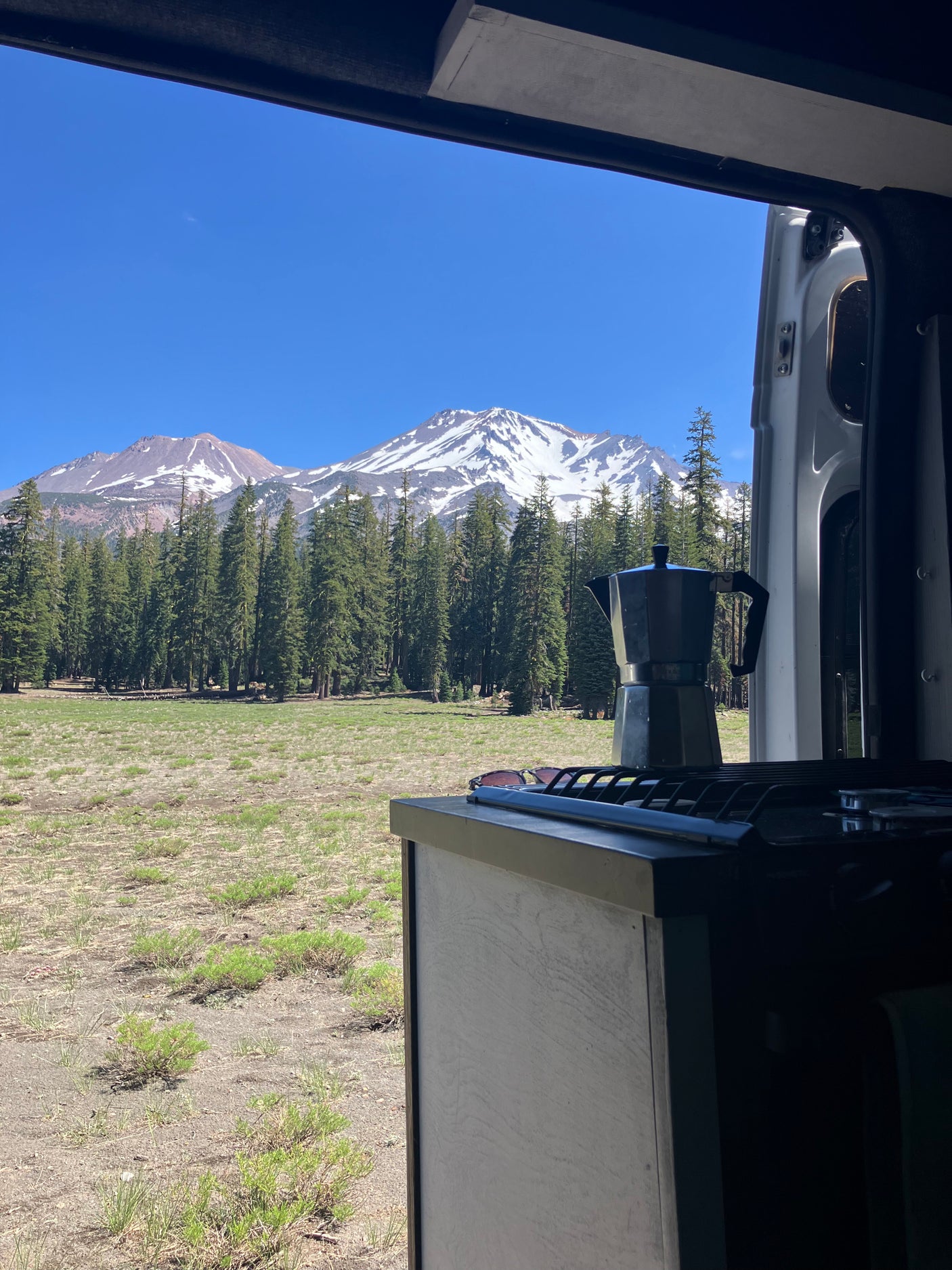 RV rental view: Moka pot on counter, looking out to a vast forest and majestic snow-capped mountains under a clear sky.