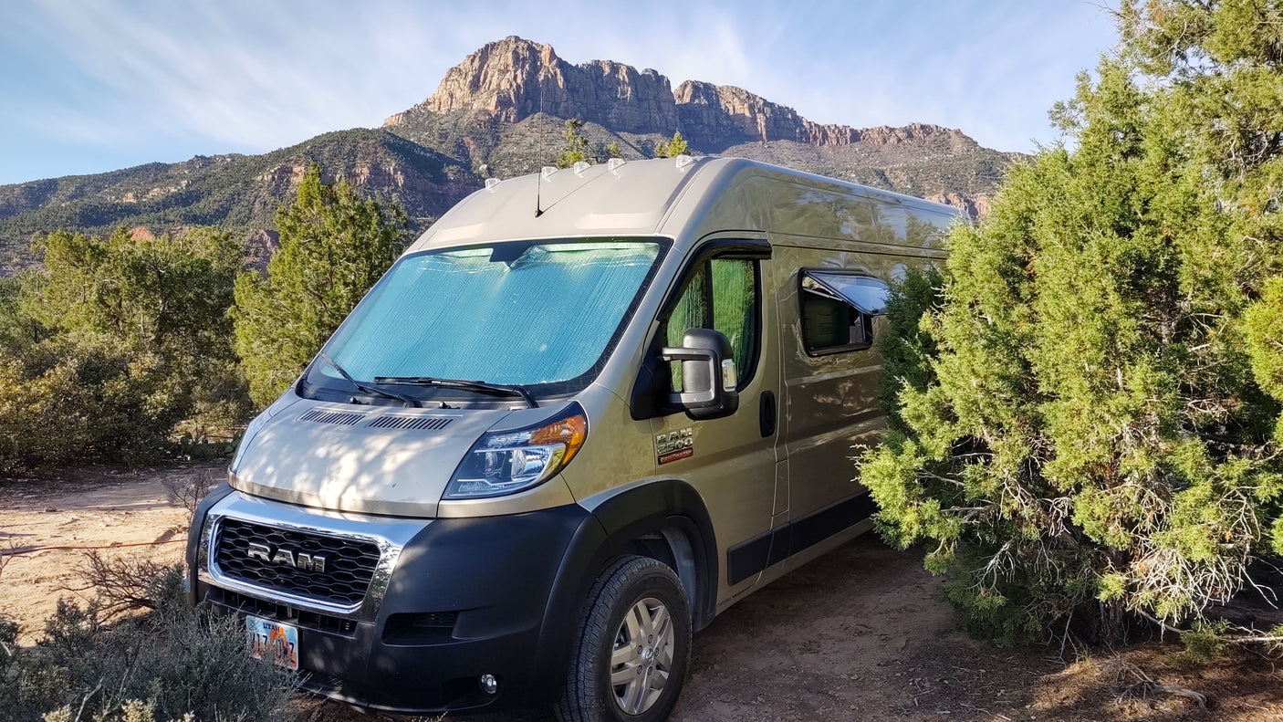 Golden Ram ProMaster RV rental van sits on a dirt path, nestled among green trees with majestic mountains in the background.