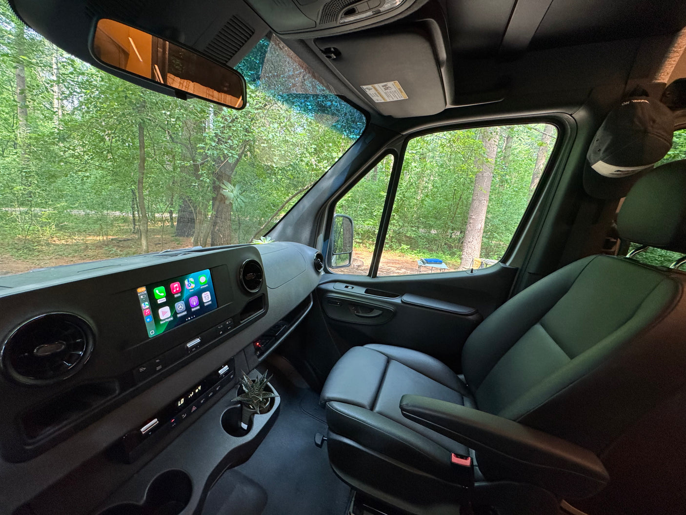 Inside an RV rental van at a forest campsite: dashboard with digital display, leather passenger seat, and outdoor table.