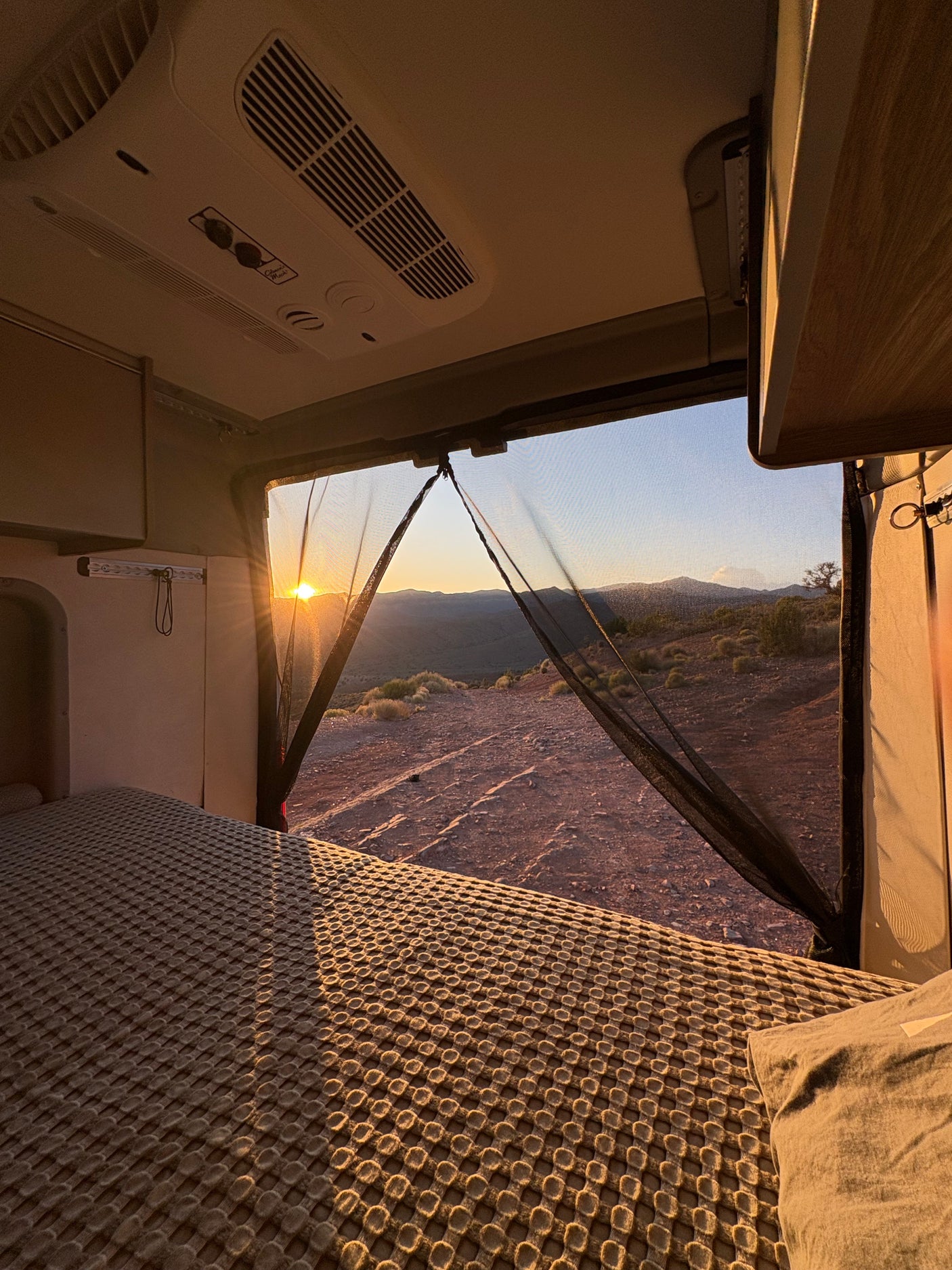 Inside an rv rental: a cozy bed overlooking a breathtaking desert sunset and mountain range. Ideal for off-grid camping.