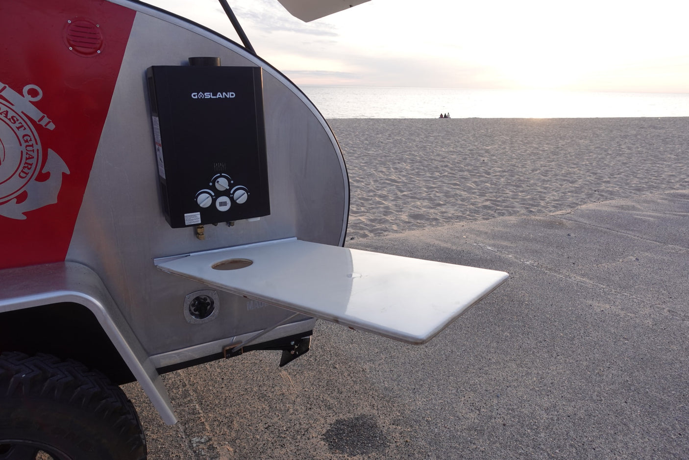 Visually, a red and silver teardrop **rv rental** is parked by a sandy beach, featuring an exterior water heater and a fold-out table.