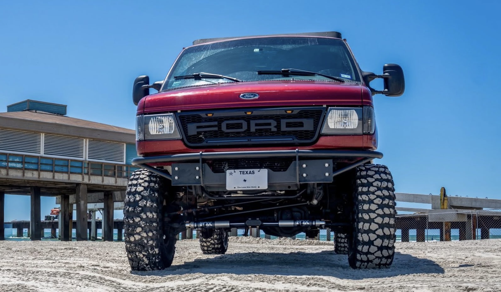 This lifted red Ford van, an ideal off-road RV rental, is parked on a Texas beach, ready for adventure.