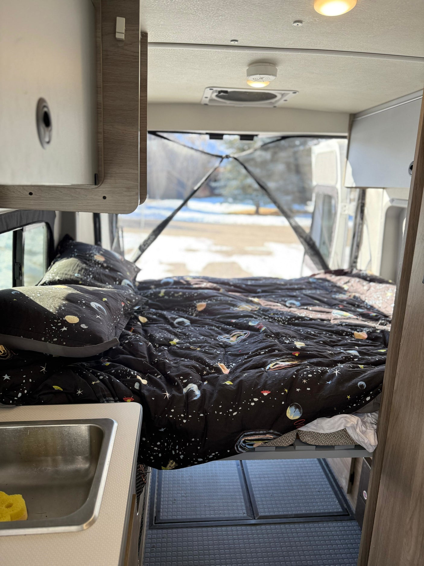 Cozy RV rental interior: space-themed bed, small sink, and a view of snow out the open rear doors.