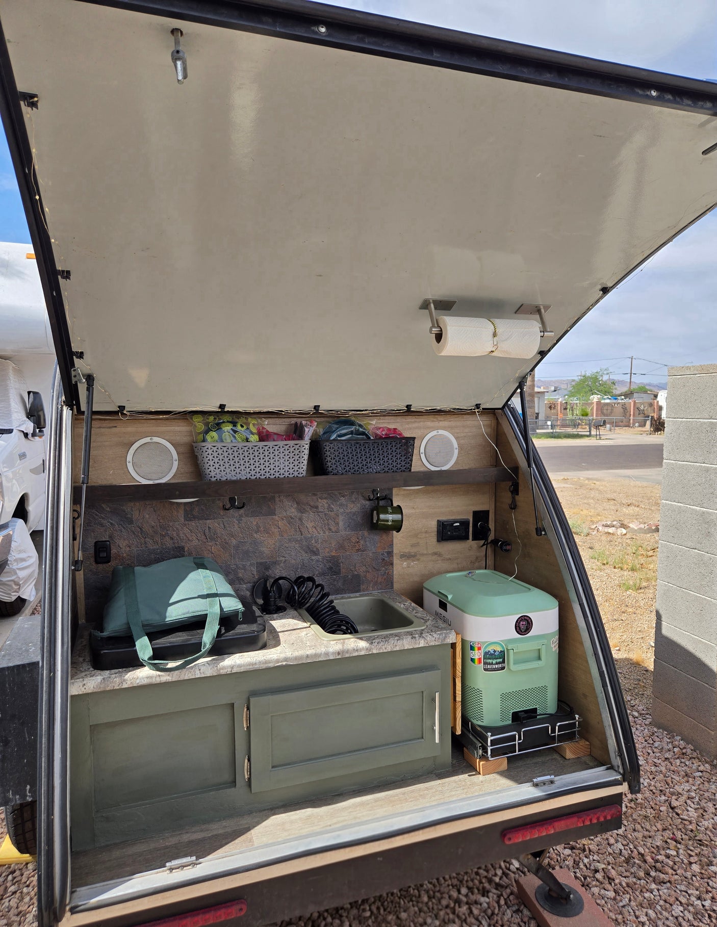Open RV rental hatch reveals a compact outdoor kitchen: sink, portable fridge, storage, speakers, and paper towels.