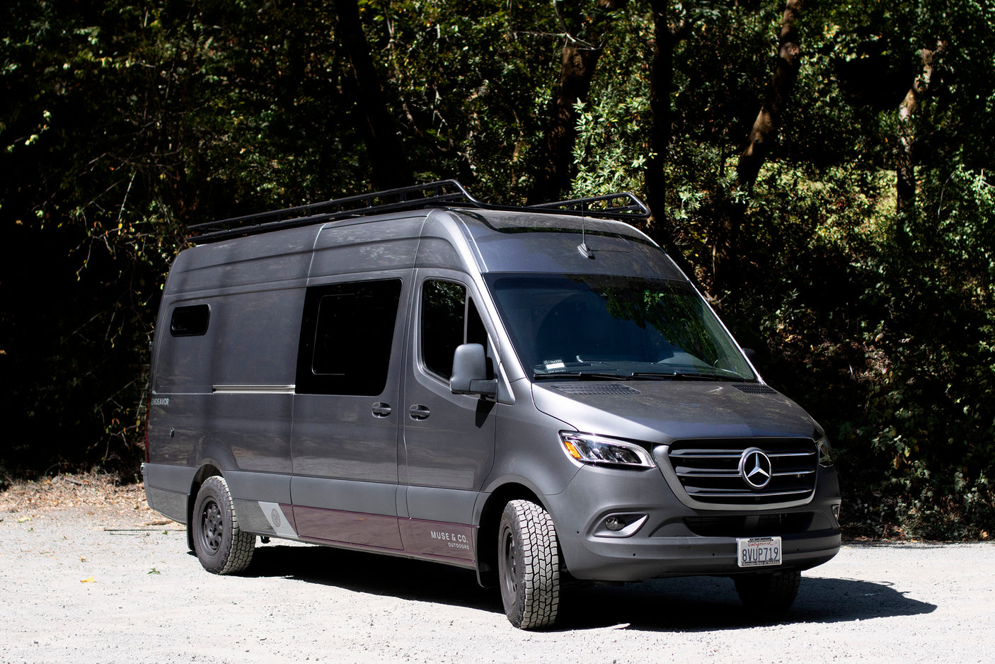 A gray Mercedes Sprinter camper van for RV rental, ready for outdoor adventure on a gravel road by lush trees.