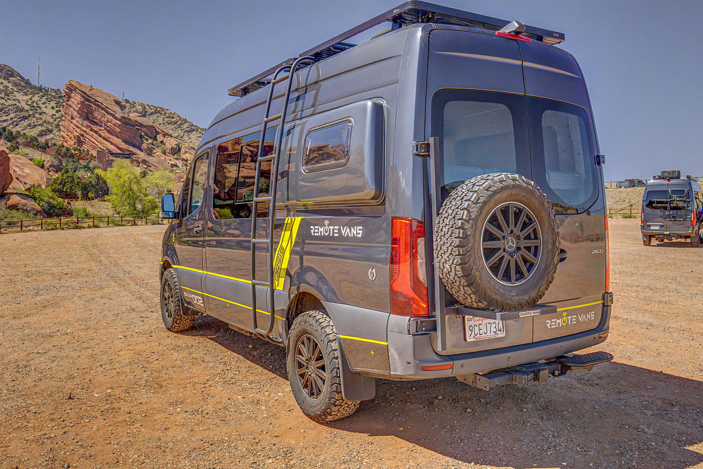 Adventure-ready gray Sprinter RV rental with off-road tires and roof rack at a scenic red rock park.