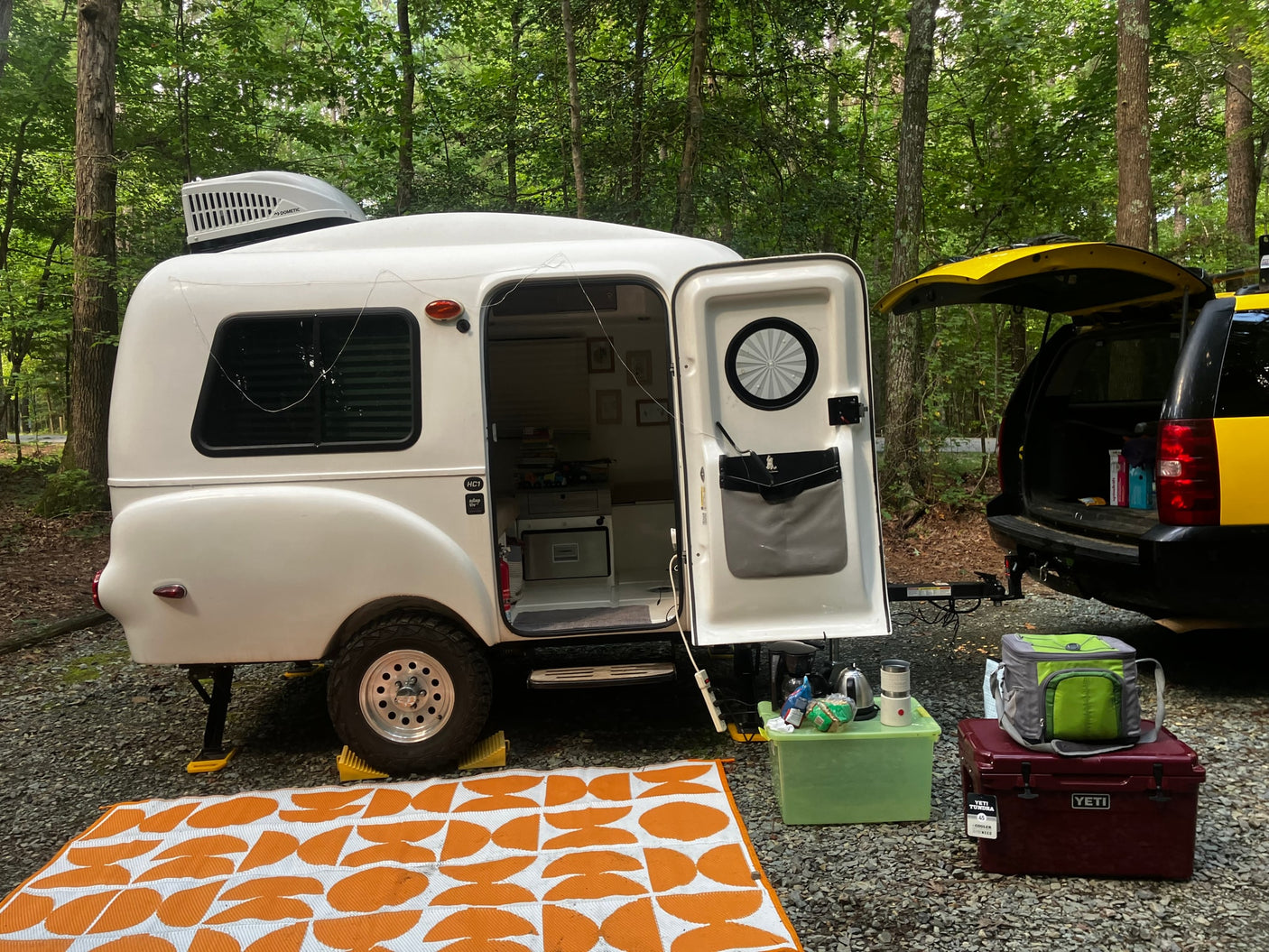 White HC1 rv rental camper & yellow SUV ready for adventure at a forest campsite. An orange mat & camping gear complete the setup.