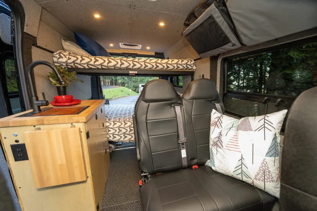 Inside an rv rental: a compact kitchen with a sink, an elevated bed, and two rear passenger seats for travel.