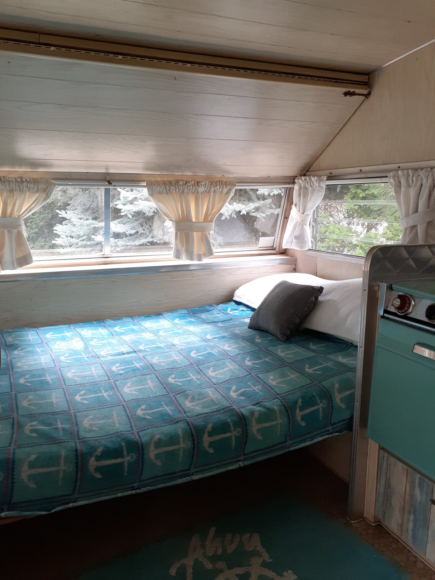 This cozy rv rental interior features a bed with an anchor blanket, bright windows showing trees, and a teal kitchen unit.