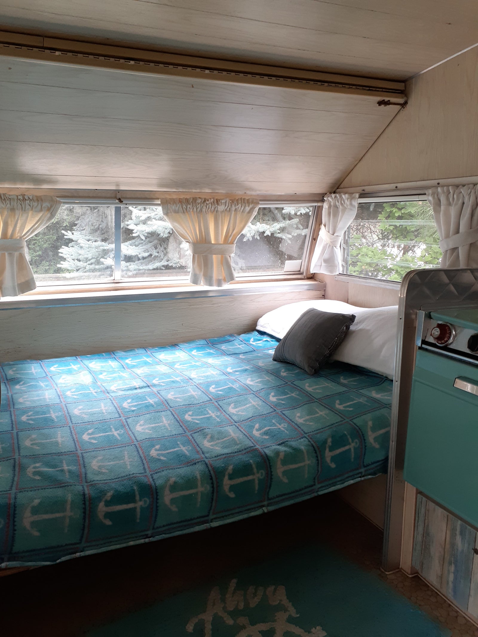 This cozy rv rental interior features a bed with an anchor blanket, bright windows showing trees, and a teal kitchen unit.