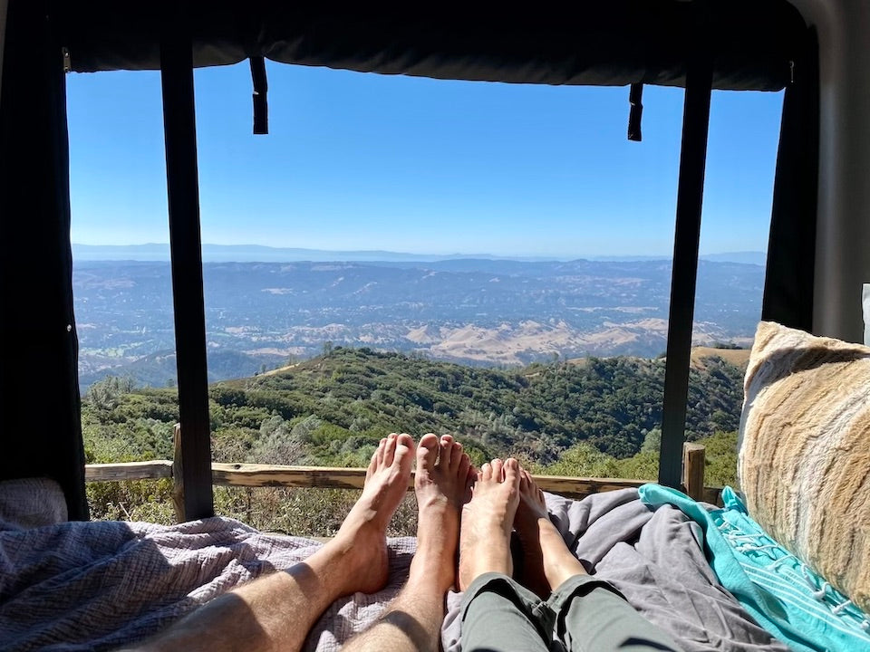 Couple's feet up, enjoying an expansive mountain and valley view from a cozy RV rental bed. Perfect escape.
