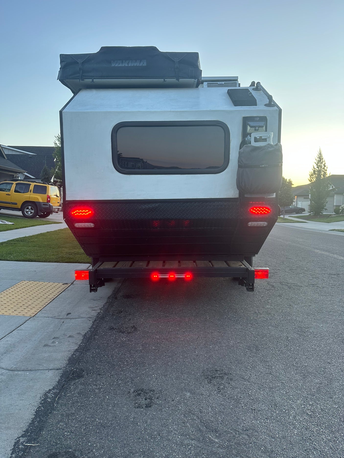 Rear view of a compact RV rental: an overland trailer with a Yakima rooftop tent, propane tank, and bright red taillights.