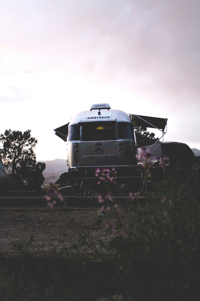 Discover adventure with an RV rental! A classic silver Airstream trailer is parked at dusk in a scenic outdoor setting, with flowers in the foreground.