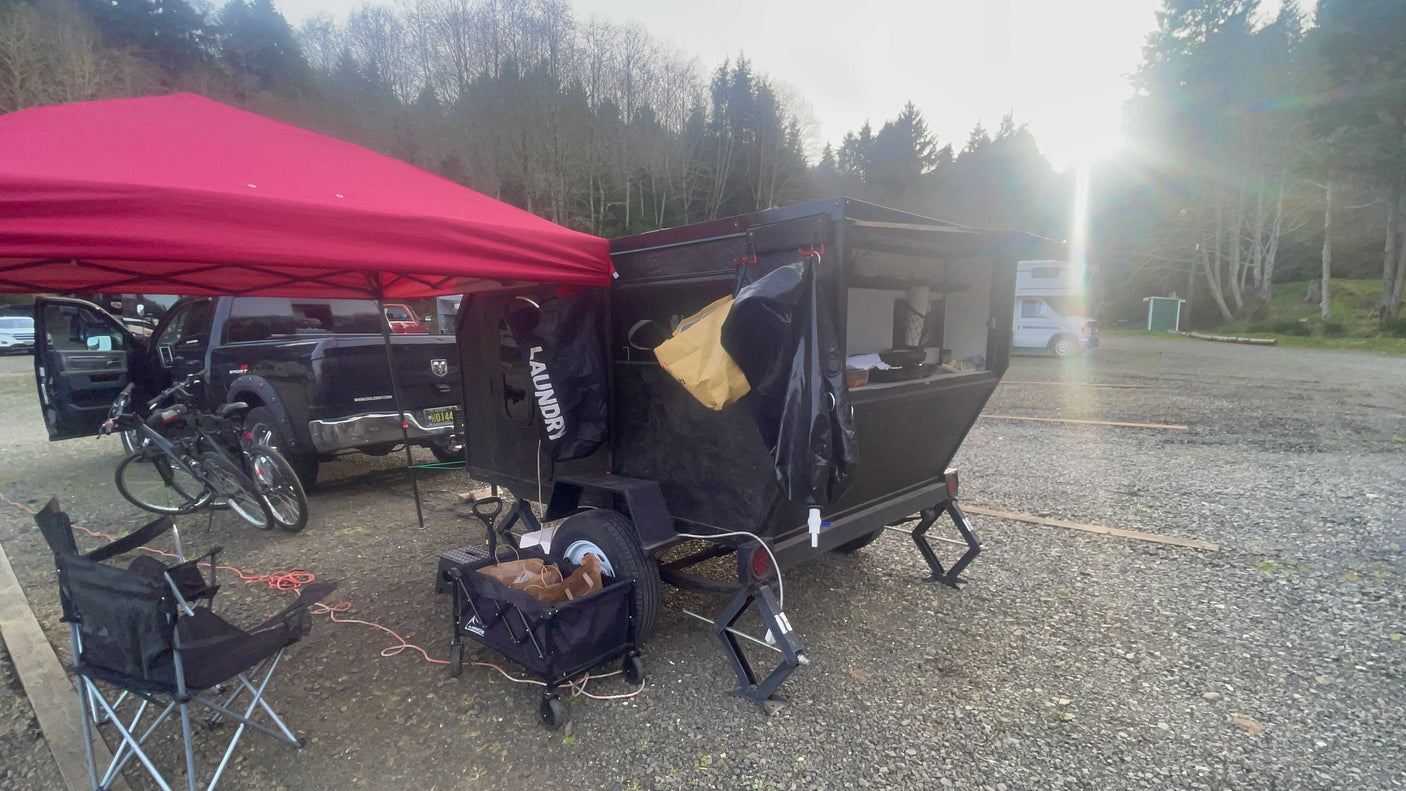 A camping setup with a red pop-up tent, truck, and custom utility trailer packed with gear, plus an RV rental in the distance.