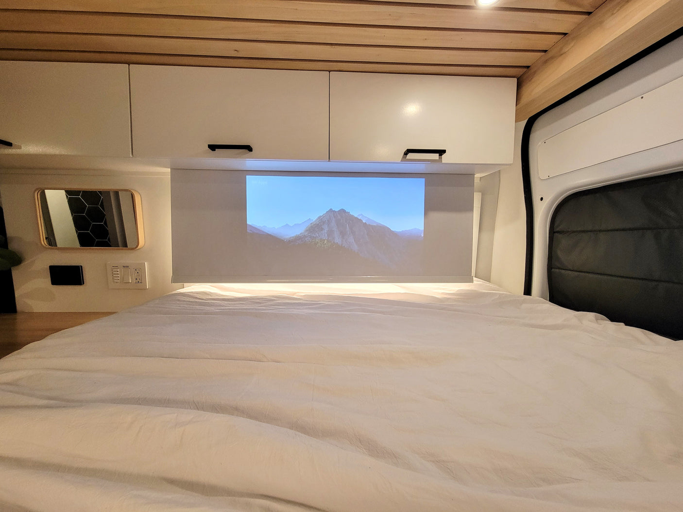 Relax in your cozy rv rental with a projector screen displaying mountains, overhead cabinets, and a comfortable bed.
