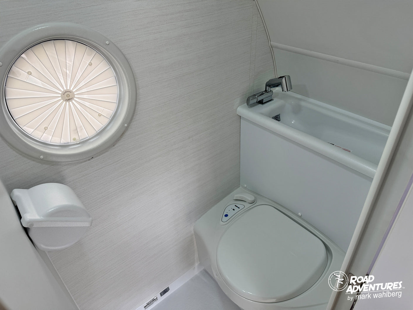 Compact RV rental bathroom: toilet, sink, and a round window with a pleated shade. Perfect for road adventures.
