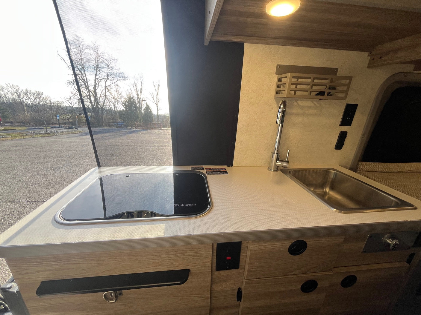 Compact RV rental kitchen with stovetop, sink, and cabinets, looking out to a sunny campground through a screen window.