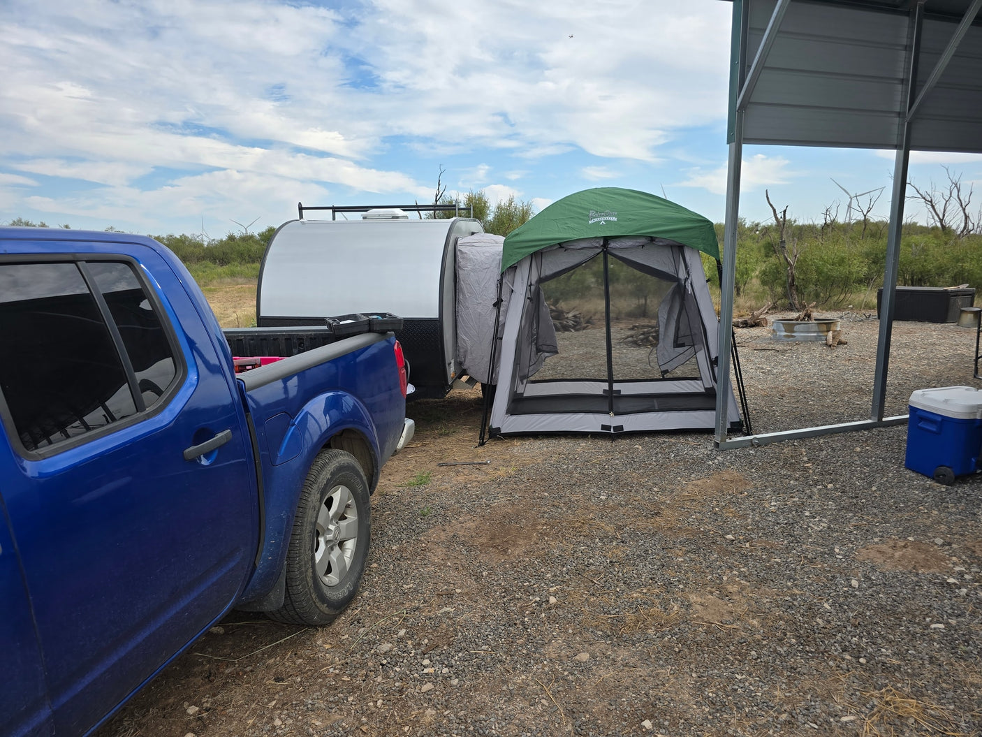 Campsite adventure with an RV rental: Blue truck, teardrop camper, & screen room. Gravel lot, arid background, wind turbines.