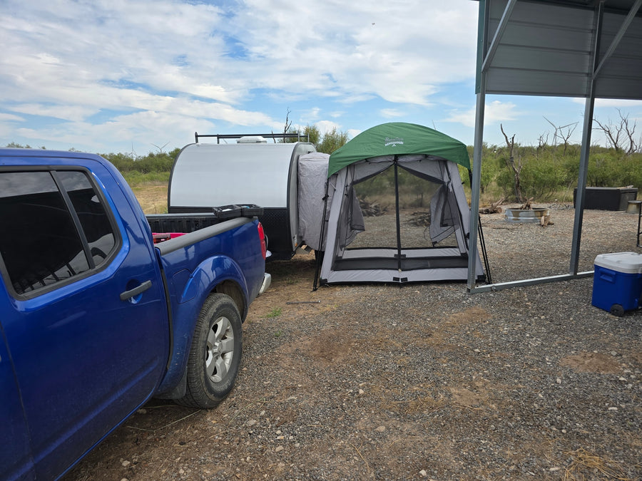 Campsite adventure with an RV rental: Blue truck, teardrop camper, & screen room. Gravel lot, arid background, wind turbines.