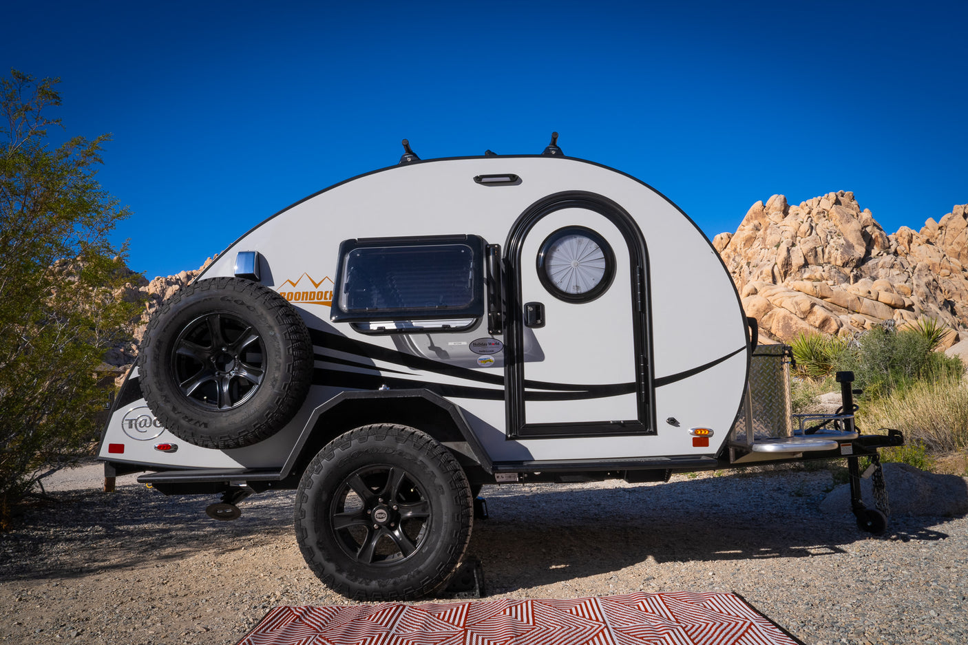 This compact teardrop RV rental, ideal for desert camping adventures, is parked on gravel with rocky terrain and a clear blue sky.