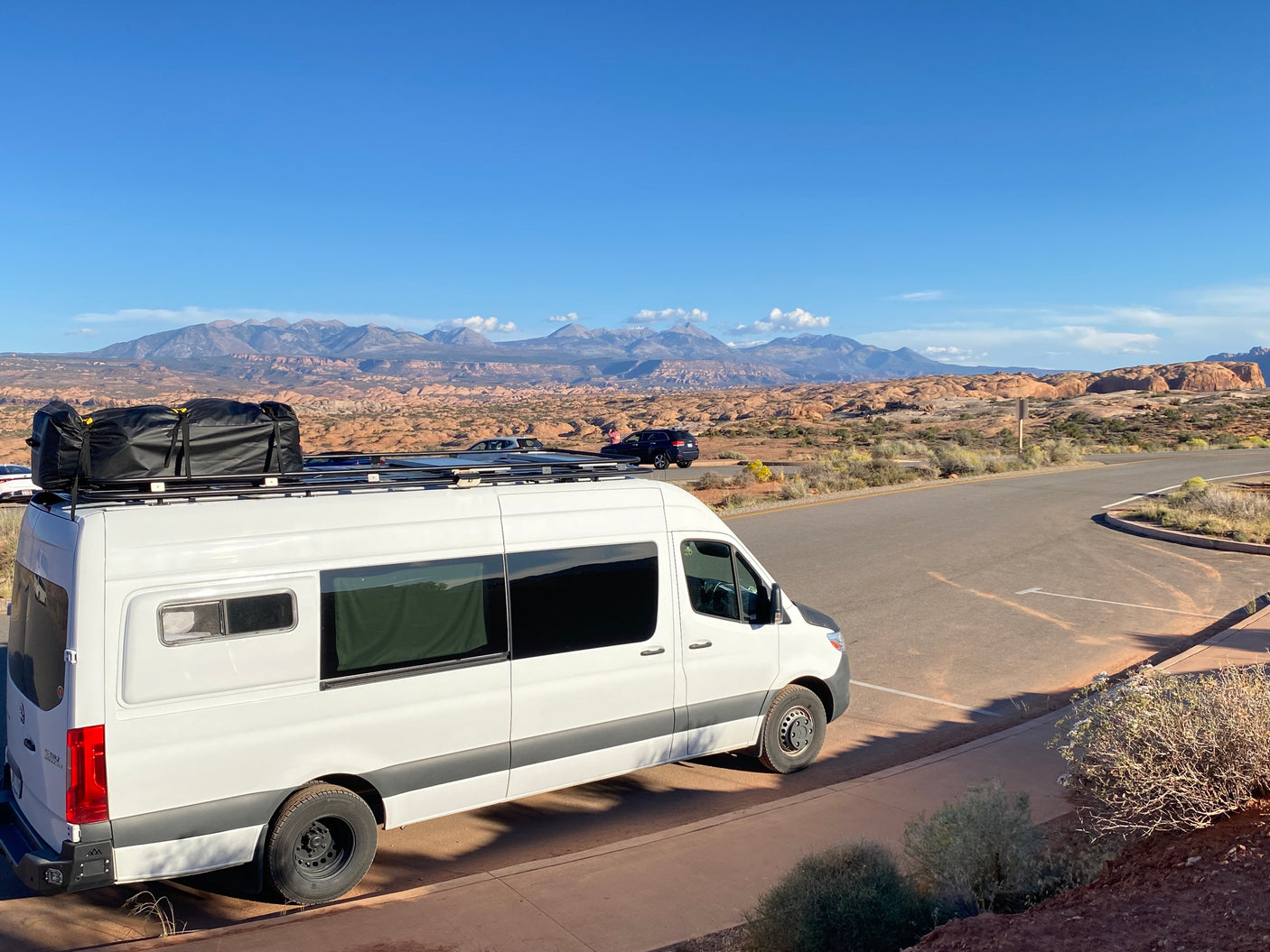 is parked against a backdrop of towering red rock formations in a sunny national park.|White rv rental Sprinter van by a calm bay