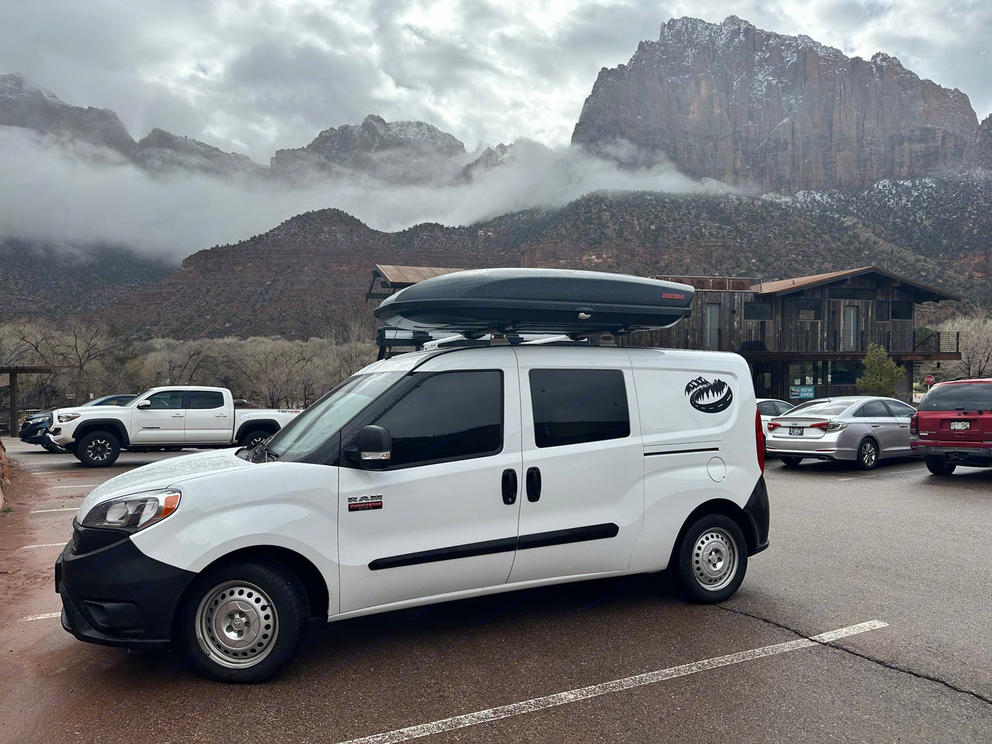 White RV rental van with Yakima roof box parked at Zion National Park, surrounded by majestic, mist-covered mountains.