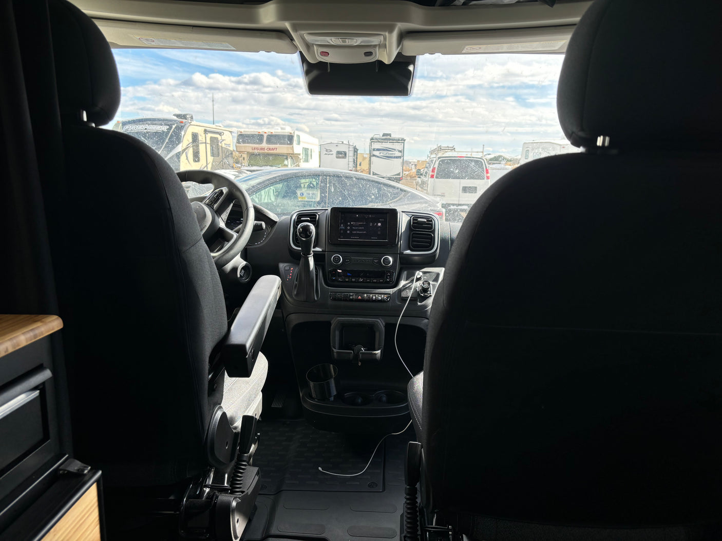 Front interior of an rv rental camper van, showing the dashboard and a view of multiple parked RVs through the windshield.