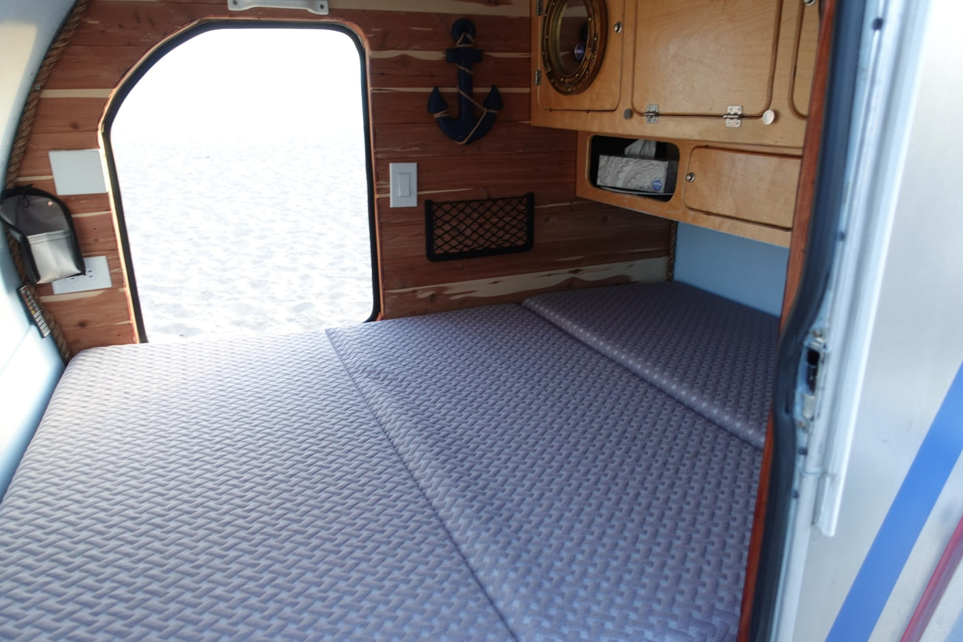 Comfortable bed in an RV rental with wood paneling, nautical decor, and an open door to a bright, sandy outdoor view.