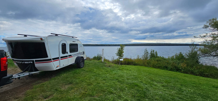 A white teardrop camper trailer (possibly an RV rental) sits on a grassy lakeside campsite under a cloudy sky.